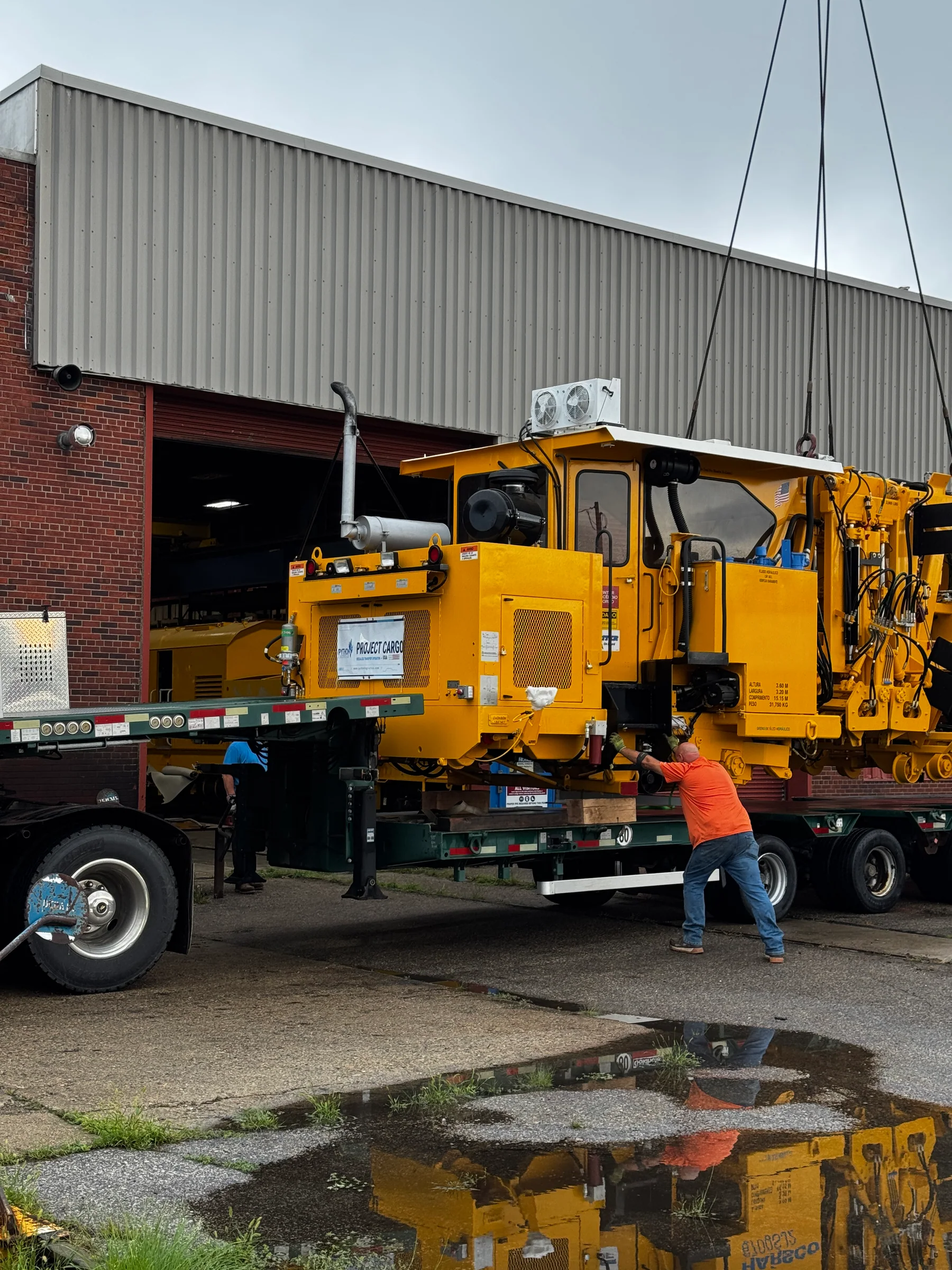 Crane slings hold yellow railway equipment above a road trailer as a worker guides the movement.