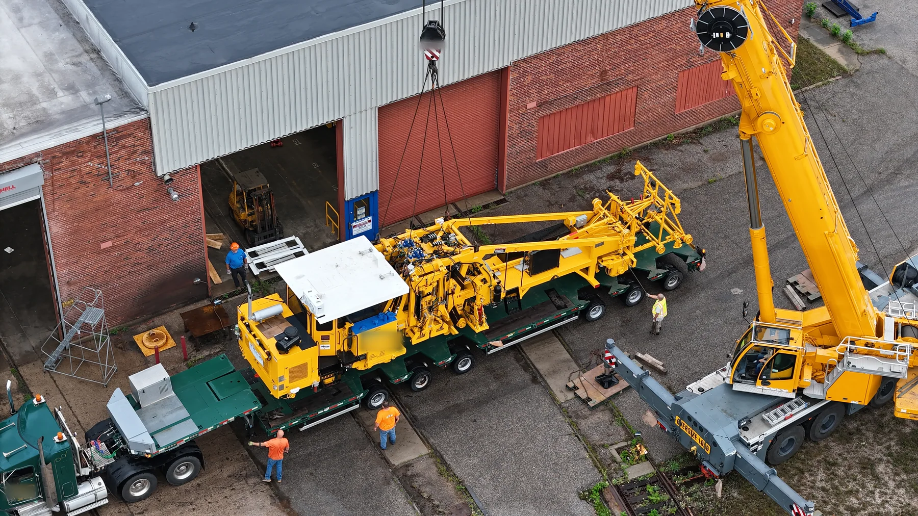 Aerial view of yellow railway equipment on a green trailer beside a crane.